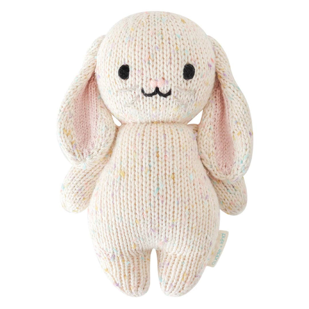 The Cuddle + Kind Baby Bunny - Confetti is a soft, hand-knit stuffed bunny toy with long, pink-accented ears, black eyes, and a sweet smile. Made from cream cotton yarn with colorful speckles—ideal for cuddling or gifting.