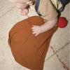 A baby in a tan shirt and denim overalls lies on a carpet, reaching for Pepa the Apple Big Doudou—an organic cotton comfort toy featuring a brown cloth with a small red and green fabric apple attached.