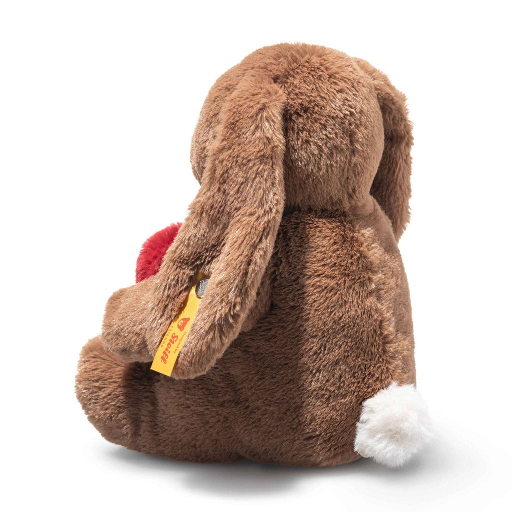 The Steiff Hoppie Rabbit – Love plush sits facing away, holding a red heart. It has a white tail, long ears with a yellow tag, and is set against a plain white background—a perfect gift for Steiff fans.