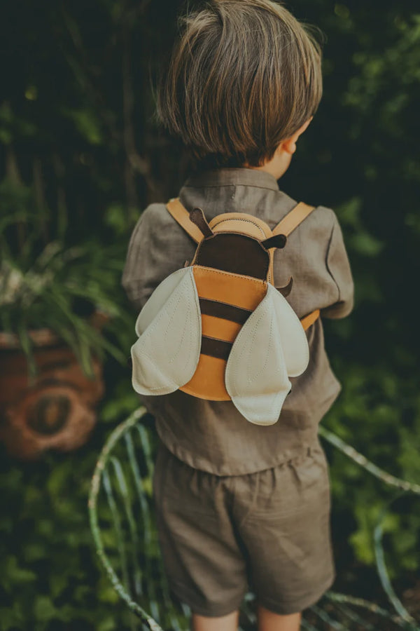 A young boy in a linen shirt and shorts, viewed from behind, wears a small Donsje Bee Backpack made of classic leather, shaped like bee wings, standing against a lush green backdrop.