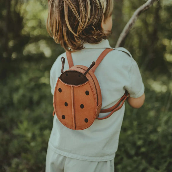 A child from behind, wearing a Donsje Mini Leather Backpack - Ladybug, stands in a sunlit forest. The child is holding a stick and dressed in a pale blue shirt.