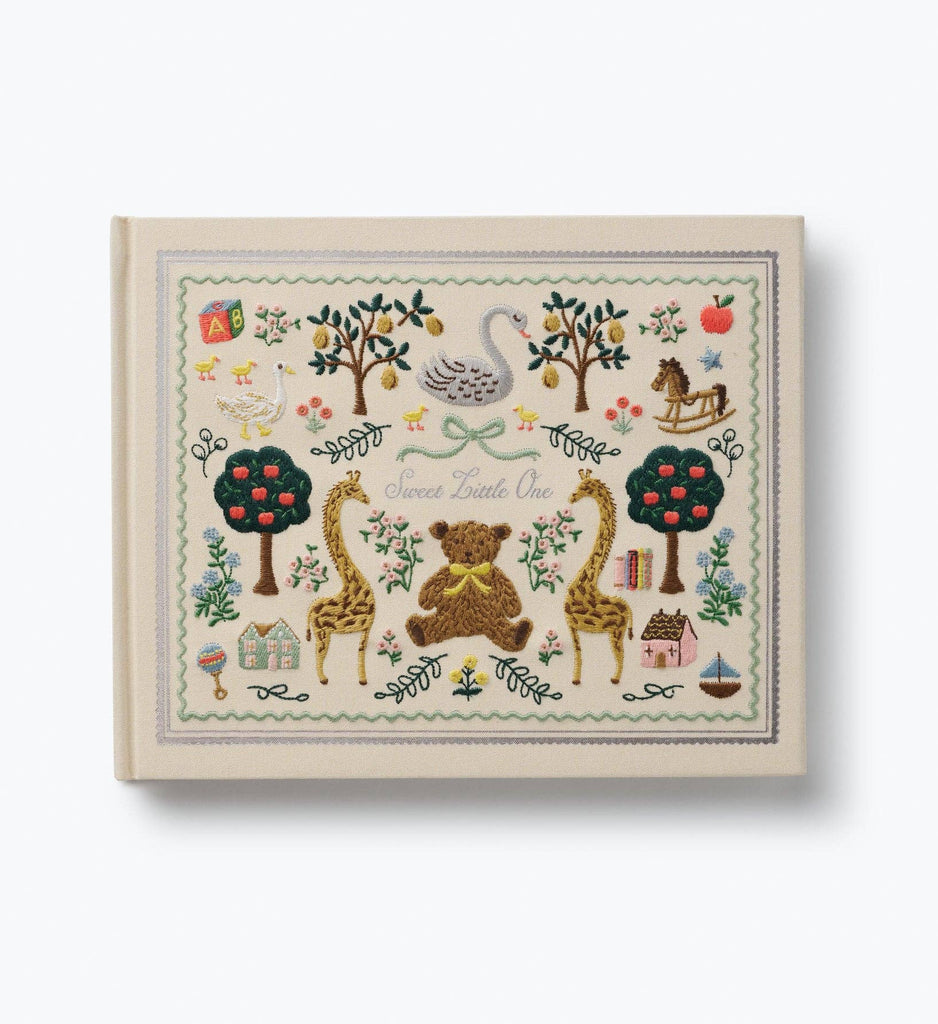 Introducing the Storybook Embroidered Baby Book: a cream-colored memory book adorned with delicate embroidery and charming illustrations of a bear, giraffes, trees, swan, rocking horse, houses, and flowers. The elegant cursive cover reads “Sweet Little One,” making it the perfect keepsake for capturing your child’s precious milestones in timeless style.