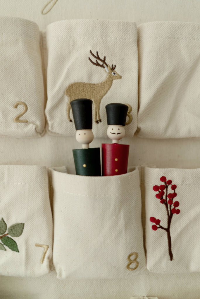 Two wooden peg dolls in black hats and painted uniforms sit in a cloth pocket marked with an 8 on the Sabo Concept Christmas Cotton Advent Calendar, which features embroidered branches and a deer on cream cotton.