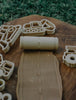 The Road Roller - Eco Pattern Playdough Roller stamps a road pattern onto brown dough, accompanied by construction vehicle-shaped cookie cutters, all arranged on a wooden board with grass in the background. This eco-friendly playdough roller is a perfect addition to any set of sensory play tools.