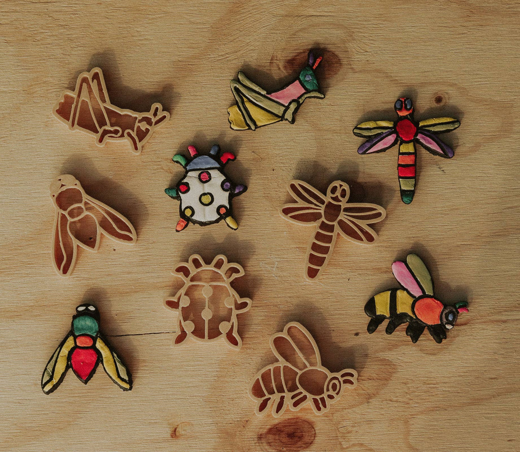Experience creative play or baking inspiration with the Insect Cutter Set - Eco Playdough Tool, featuring assorted decorated and undecorated insect-shaped cookies—dragonflies, bees, ladybugs, and a grasshopper—arranged on a wooden surface. Perfect for your next baking session or sensory play with this eco-friendly set.