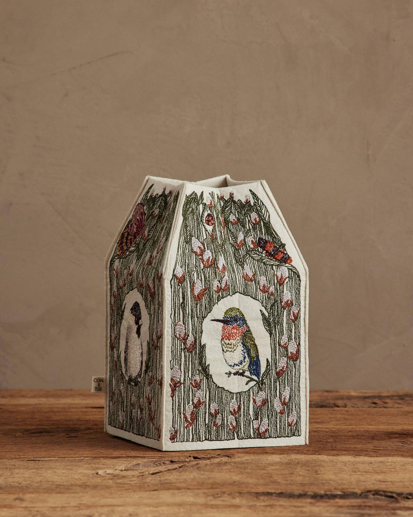 The Coral & Tusk Birdhouse Tissue Box Cover, featuring embroidered birds and floral designs, is displayed on a wooden surface against a beige wall.