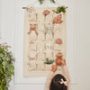 A child reaches for the Cuddle + Kind Baby Snowman - Candy Cane from a cream-colored fabric advent calendar with numbered pockets, each holding hand-knit baby animals and festive décor made from Peruvian alpaca yarn, hanging on a wall by greenery.
