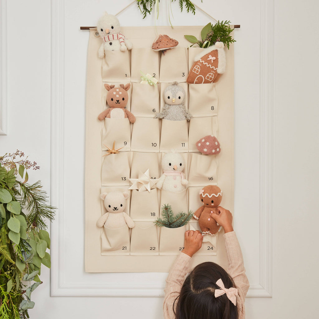 A child reaches for the Cuddle + Kind Baby Snowman - Candy Cane from a cream-colored fabric advent calendar with numbered pockets, each holding hand-knit baby animals and festive décor made from Peruvian alpaca yarn, hanging on a wall by greenery.