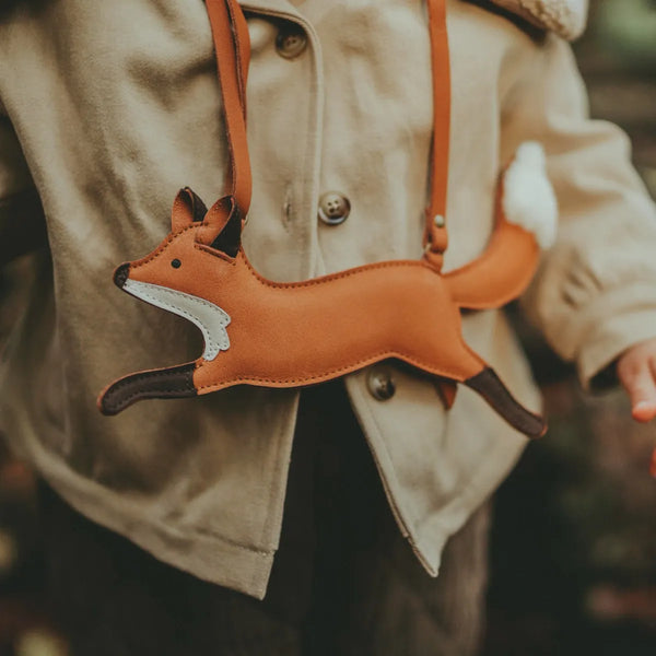 A child wearing a beige coat carries a Donsje Jimp Purse - Fox with a white tail, suspended by adjustable leather straps. The background is softly blurred, highlighting the vibrant orange of the fox.