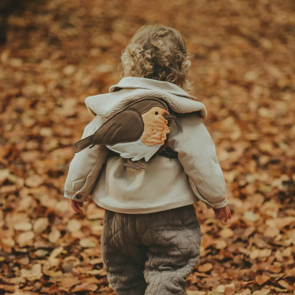 A toddler with curly hair wearing a Donsje Woodsy Backpack - Robin walks on a leaf-covered path in autumn.