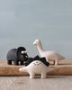 Three handcrafted wooden animal figurines—a black elephant, a white Brachiosaurus, and a cream swan—arranged on a wooden ledge against a plain backdrop.