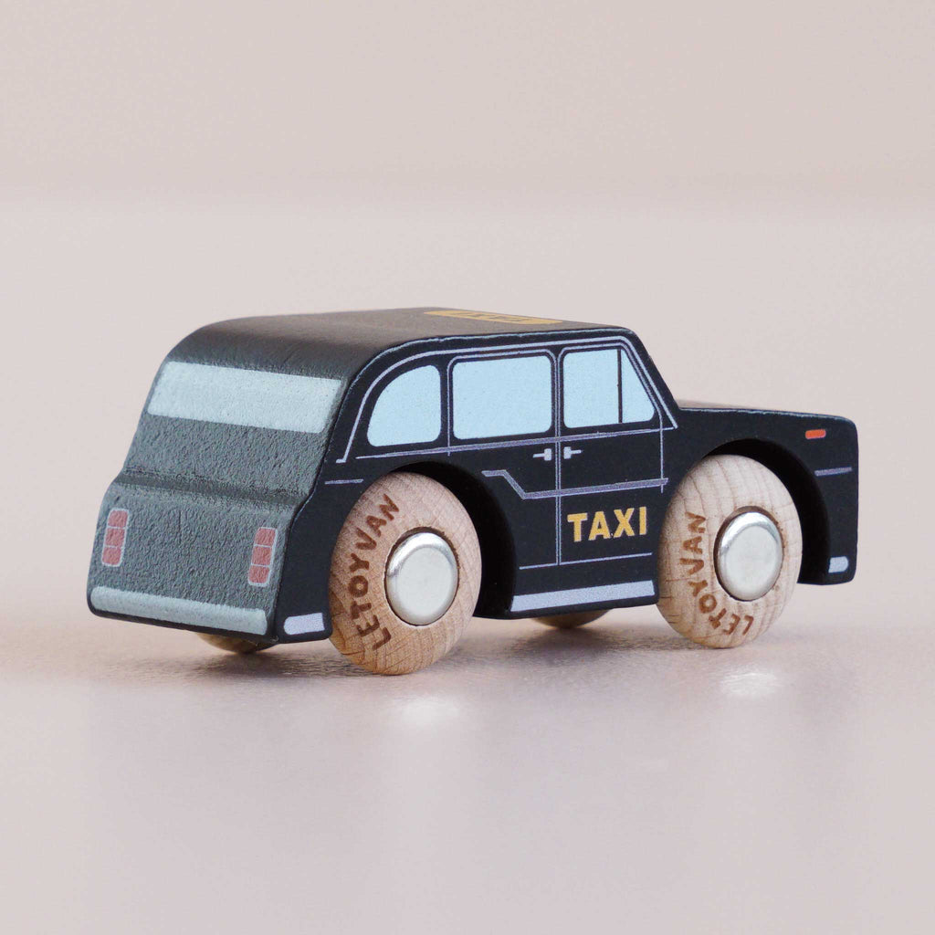 A small wooden toy taxi, the No. 45 GT275 Bertie made in England, styled as a classic London black cab with “TAXI” written on the side, sits on a light-colored surface. The plain wooden wheels feature “LETOYVAN” printed on them.
