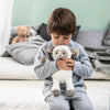 A young boy in a gray sweater hugs a Lita Lamb Plush Animal Toy while sitting on the floor of a room with soft colors and a blurred background of teddy bears and cushions.
