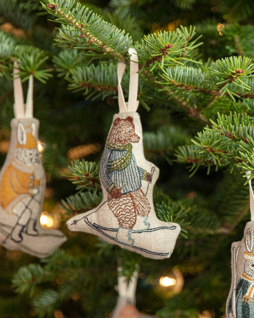 The Coral & Tusk Downhill Bear Ornament, featuring a bear in a scarf and sweater on skis, hangs from a green pine Christmas tree with softly glowing lights in the background.