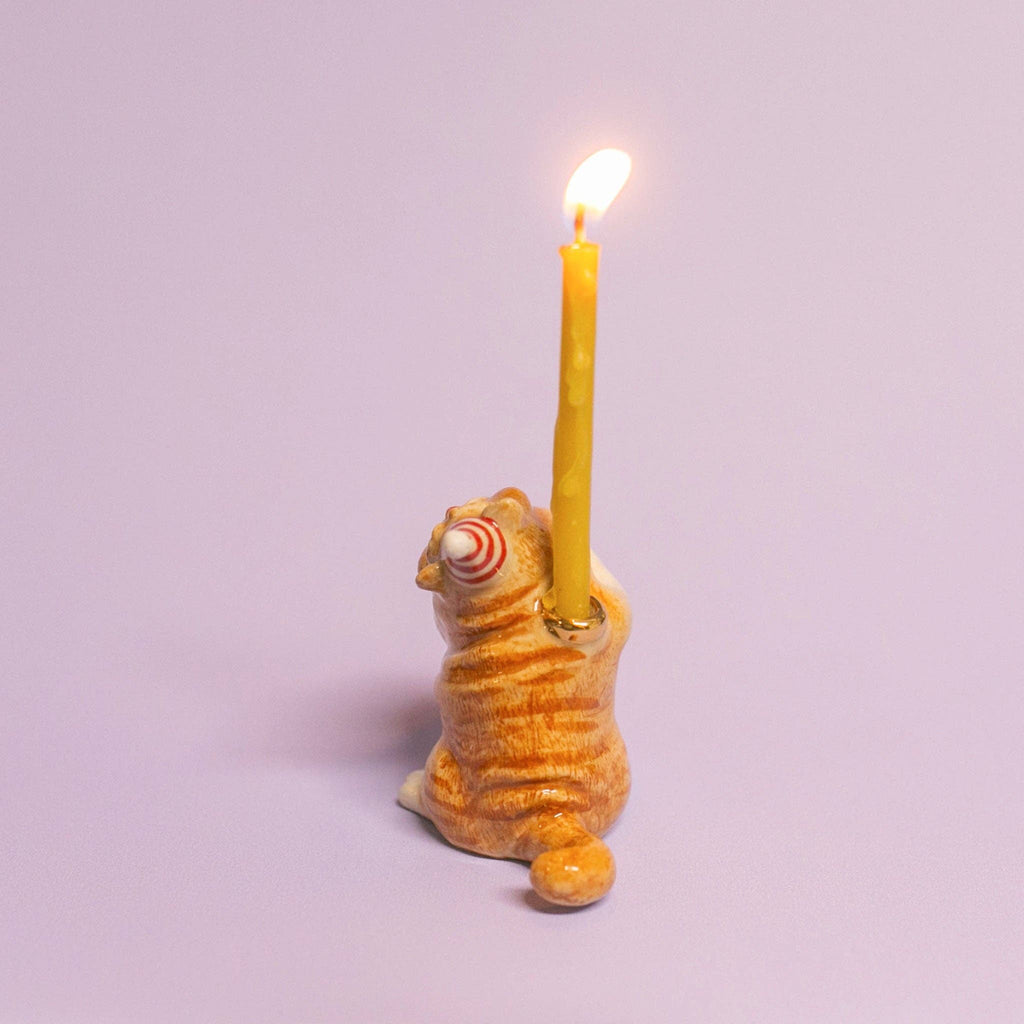 The Exotic Shorthair Cat Cake Topper features a ceramic cat with orange stripes, back turned and holding a lit yellow candle. Ideal for cat-themed parties or decor, it pops against a plain light purple background.