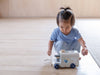 A young child in a blue outfit sits on a light wooden floor, playing with the Sorting Bus - Orchard Series, a white pull toy train with blue wheels and a pull string, reminiscent of classic shape sorter toys.