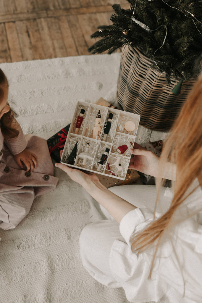 A person holds a box of Sabo Concept Christmas Ornaments beside a small child in a pink dress, with a decorated Christmas tree in a wicker basket on a textured white surface.