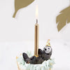 A birthday cupcake adorned with light blue frosting and featuring a Sea Otter Cake Topper wearing a party hat and holding a lit gold candle.