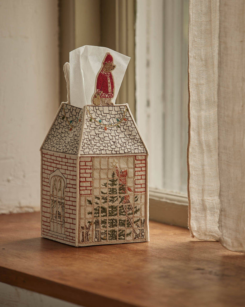 The Coral & Tusk Home for the Holidays Tissue Box Cover, shaped like a house with festive embroidery, features a Christmas tree, windows, and a person in winter clothing holding a tissue on the windowsill.