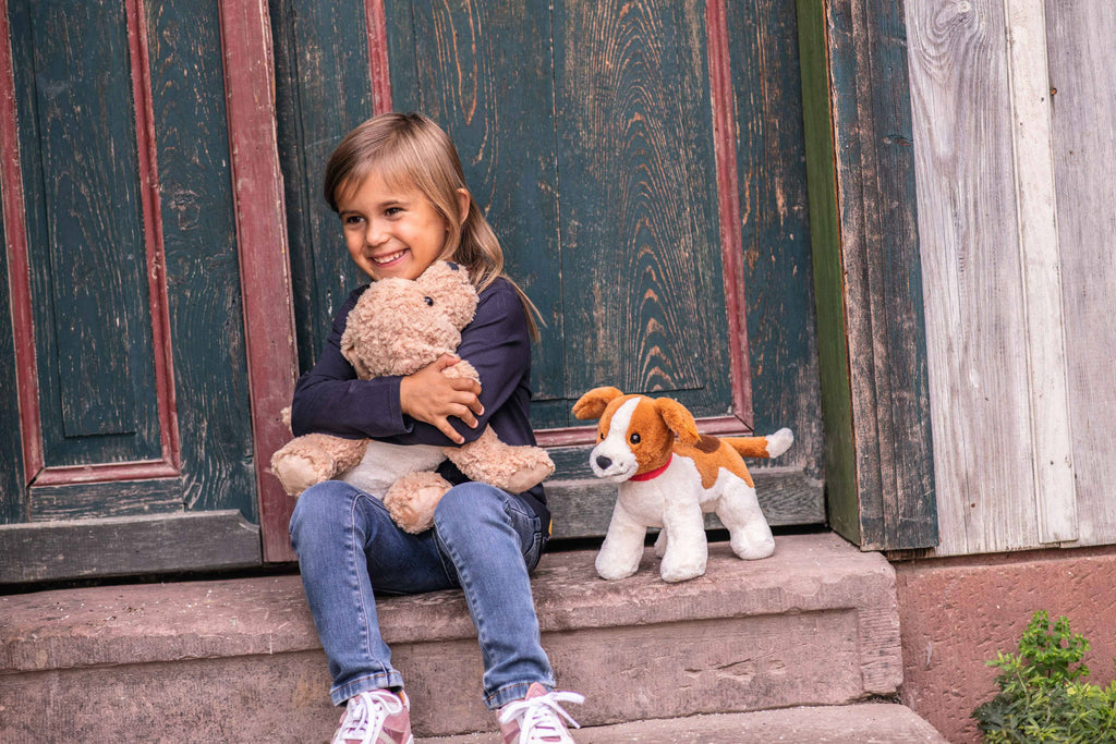 A smiling young girl sits on a step in front of a rustic wooden door, hugging her teddy bear. Beside her rests a Steiff Berno Goldendoodle Dog Plush Stuffed Animal. She's dressed in a dark long-sleeve shirt, jeans, and sneakers.