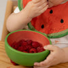 A young child uses the Wally the Watermelon Bowl & Spoon Set, enjoying fresh raspberries. Raspberry juice stains the watermelon-themed bib and table, and the wooden baby spoon from the set rests nearby as they eat.