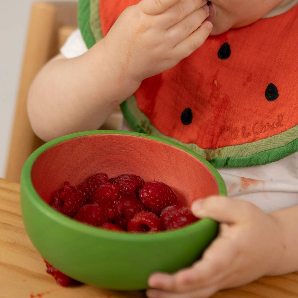 A young child uses the Wally the Watermelon Bowl & Spoon Set, enjoying fresh raspberries. Raspberry juice stains the watermelon-themed bib and table, and the wooden baby spoon from the set rests nearby as they eat.