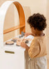 A young child stands at a wooden play kitchen, holding a spoon and stirring in a toy pot from their Wooden Pots and Pans Playset (6 Pcs), fully engaged in pretend cooking with their favorite wooden cooking toys.