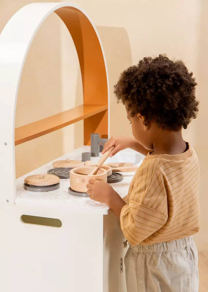 A young child stands at a wooden play kitchen, holding a spoon and stirring in a toy pot from their Wooden Pots and Pans Playset (6 Pcs), fully engaged in pretend cooking with their favorite wooden cooking toys.