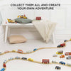The Emergency Helicopter & Rescue Cars wooden toy set, featuring colorful rescue vehicles, is arranged in a wavy pattern on the floor. A bench with a pillow, cushion, and blanket sits behind. Text: "COLLECT THEM ALL AND CREATE YOUR OWN ADVENTURE.