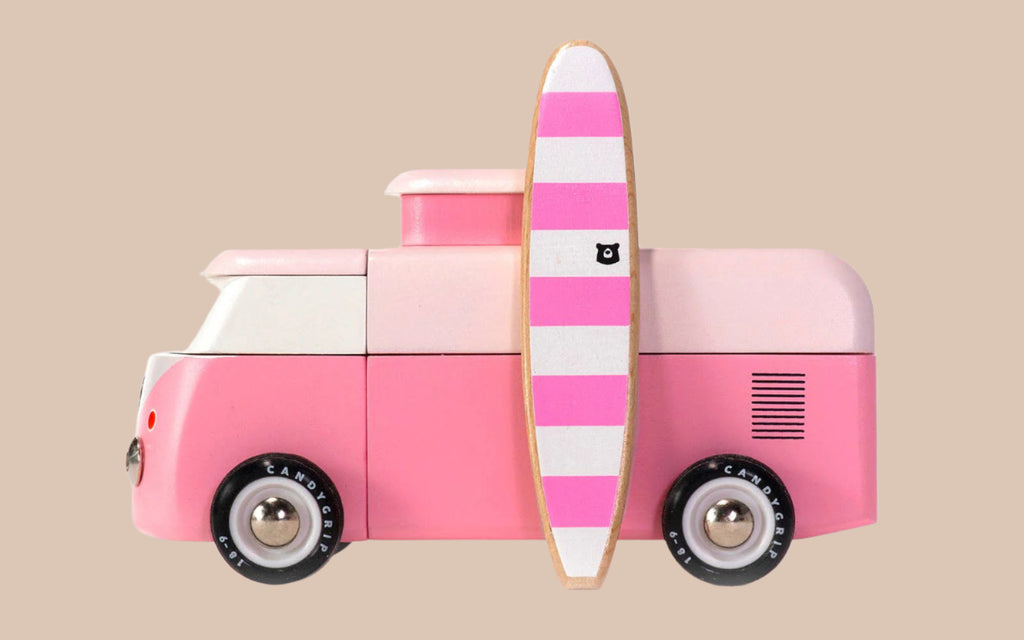 The VW Beach Bus Pink is shown in profile, featuring white accents, black wheels, and a pink-and-white striped surfboard with a small black logo mounted on the side. The background is beige.