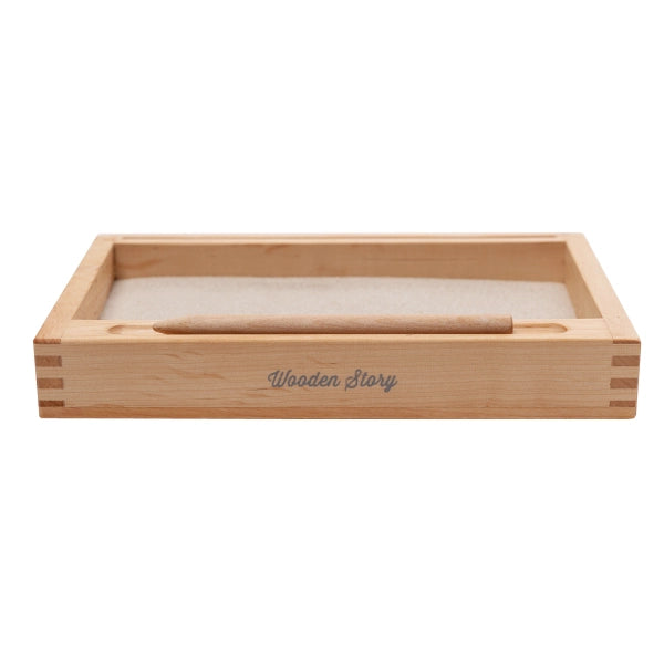 The Sand Tray with Flash-Card Holder is a rectangular wooden box with finger joints at the corners, reminiscent of a Montessori Tray. It features a smooth beige surface and includes a cylindrical wooden tool or stick resting within. The front of the box has the text "Wooden Story" engraved on it.