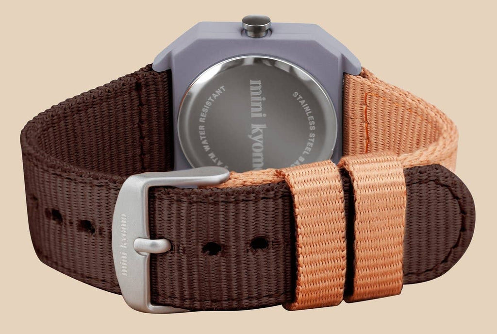 A close-up of the back of a Mini Kyomo Watch - Plum Cake with a two-tone brown and tan fabric strap, stainless steel engraved back, and a silver buckle.