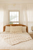 A cozy, sunlit bedroom features a wooden headboard, a neatly made bed topped with the Organic Cotton Layered Gauze Baby Blanket - Teddy (ships in one week), cream quilt, decorative pillows, white curtains, and a window letting in natural light.