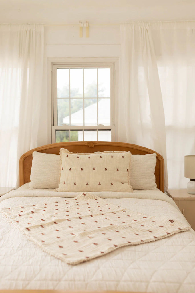 A cozy, sunlit bedroom features a wooden headboard, a neatly made bed topped with the Organic Cotton Layered Gauze Baby Blanket - Teddy (ships in one week), cream quilt, decorative pillows, white curtains, and a window letting in natural light.
