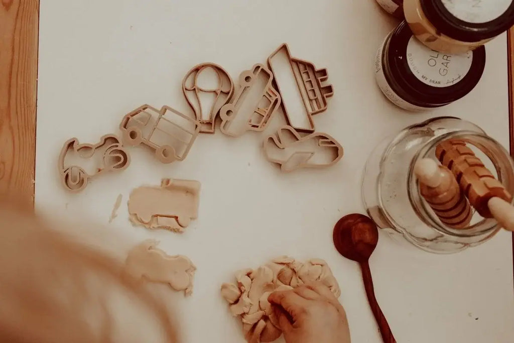 A child presses dough on a table using the Transport Cutter Set - Eco Playdough Tool, crafted from biodegradable PLA, with other Eco Playdough tools like a honey dipper, wooden spoon, and jars placed nearby.