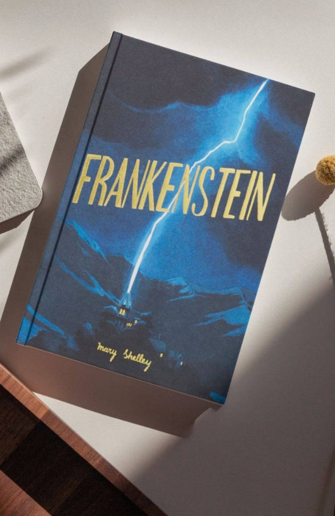 A copy of "Frankenstein" (Wordsworth Collector's Edition, Hardcover) rests on a table; its dark blue cover features a vivid lightning bolt striking a house nestled in the mountains.