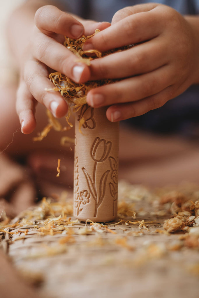 Two hands hold the Flower Roller - Eco Pattern Playdough Roller, featuring carved floral designs, while pressing dried flower petals onto its surface. Perfect for sensory play, additional petals are scattered across a woven mat below, creating an inviting scene for eco-friendly playdough fun.