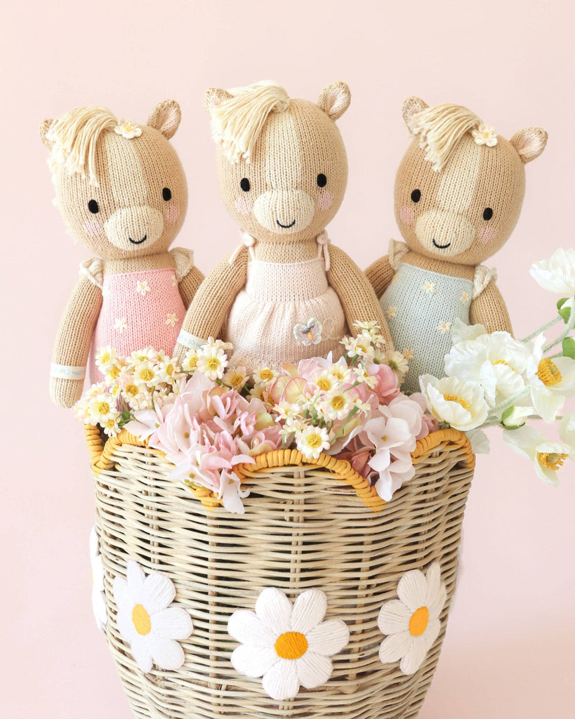 Three Cuddle + Kind Sophia the Pony dolls, handmade in Peru, wear pastel dresses as they sit in a wicker basket with white daisies, surrounded by pink and white flowers on a light pink background.