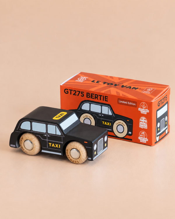 A black cab toy with wooden wheels is placed in front of an orange box labeled “No. 45 GT275 Bertie | Made in England.” The box features images and details about the London toy car.