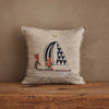 The Coral & Tusk Sailing Foxes Pillow, in beige, displays two foxes sailing with a blue-and-white patterned sail and sits on a wooden surface against a plain brown wall for a nautical accent.