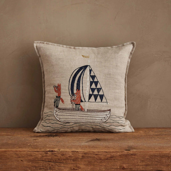 The Coral & Tusk Sailing Foxes Pillow, in beige, displays two foxes sailing with a blue-and-white patterned sail and sits on a wooden surface against a plain brown wall for a nautical accent.