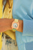 Someone wears the Mini Kyomo Watch - Watercolor in pastel yellow and green with a hexagonal peach face, paired with a light blue jacket and yellow top against a yellow background.