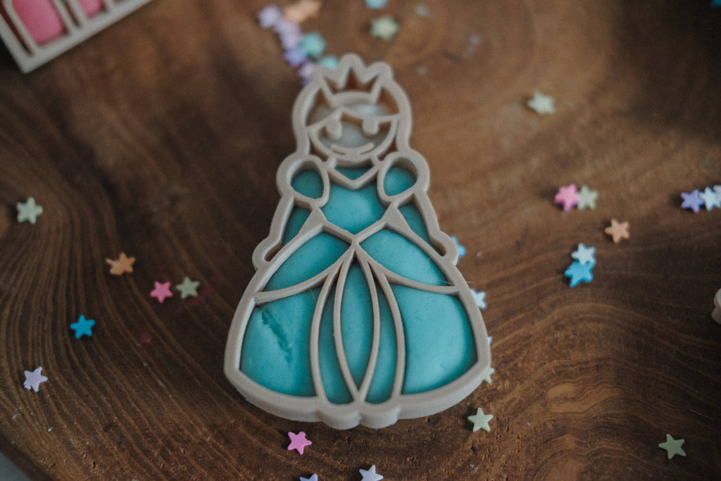 A blue princess-shaped cookie with a crown rests on a wooden surface, surrounded by small, colorful star-shaped sprinkles—expertly made with our Princess Cutter Set - Eco Playdough Tool from the princess themed playdough tools collection.