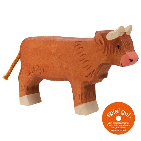 A small, handcrafted wooden toy cow with a brown body, beige horns, and a short, orange tail. The cow is standing upright and facing slightly to the right. An orange circle with white text near the bottom right indicates a "spiel gut" recommendation. This high-quality piece is part of the Holztiger Highland Cattle, Standing collection.