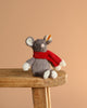 A cuddly Steiff Cozy Christmas Mia Mouse with white paws and ears, wearing a red scarf, sits on a wooden stool against a beige background—meet your festive stuffed animal friend.