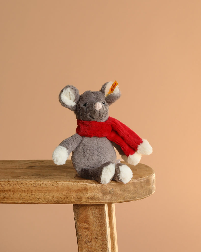 A cuddly Steiff Cozy Christmas Mia Mouse with white paws and ears, wearing a red scarf, sits on a wooden stool against a beige background—meet your festive stuffed animal friend.