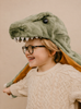 A smiling, bespectacled child in a cream sweater playfully wears the Costume - T-Rex Dino, a large plush dinosaur hat covering their head and shoulders.