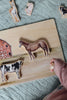 A child’s hand is holding a piece of the Farm Animals Wooden Puzzle: A Tabitha Paige Puzzle, with horse, cow, llama, sheep, and goat-shaped pieces resting on a quilted surface as they engage with this toddler educational toy.