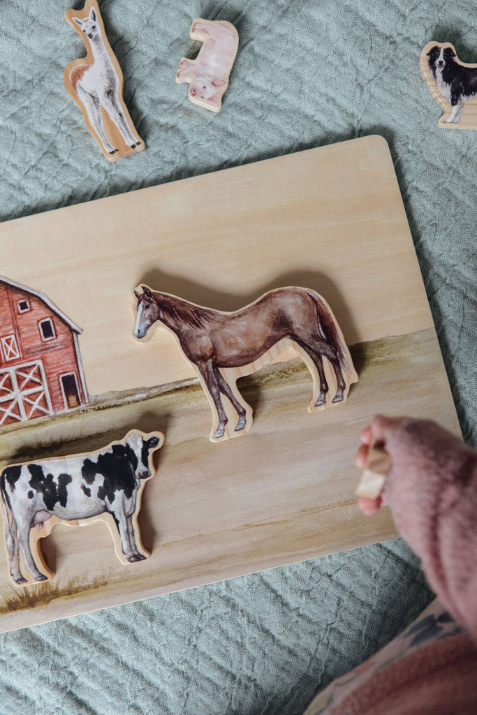 A child’s hand is holding a piece of the Farm Animals Wooden Puzzle: A Tabitha Paige Puzzle, with horse, cow, llama, sheep, and goat-shaped pieces resting on a quilted surface as they engage with this toddler educational toy.