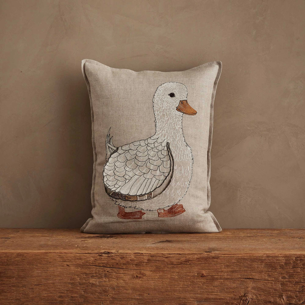 The Coral & Tusk Mama Duck Pocket Pillow features an embroidered white duck with orange beak and feet on beige fabric, displayed against a plain brown wall on a rustic wooden surface.